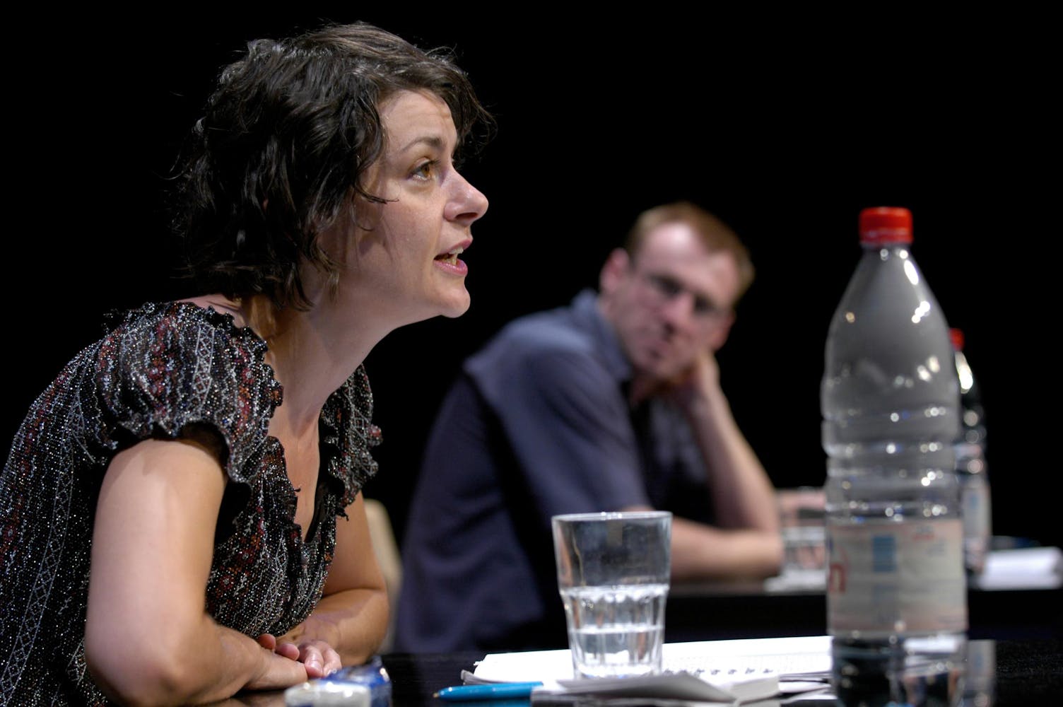 One performer with light skin speaks while another looks on.