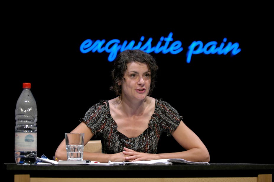 A performer with light skin sits at a desk in front of a neon sign that reads "exquisite pain".