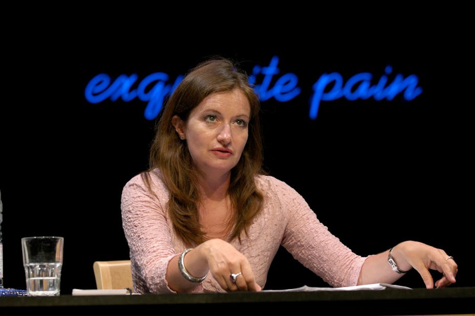 A performer with light skin sits at a desk in front of a neon sign that reads "exquisite pain".