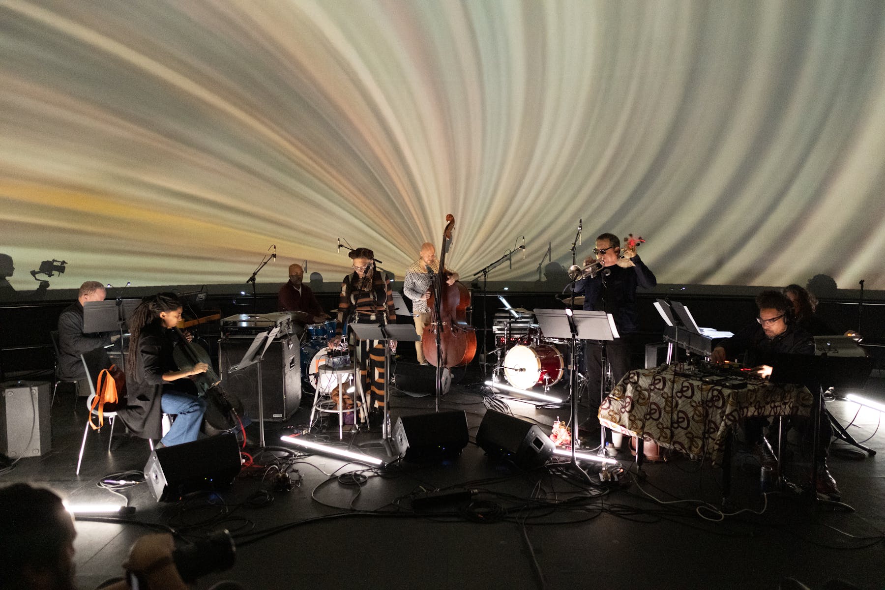 Nine musicians perform on a stage in front of a multicolored video projection.