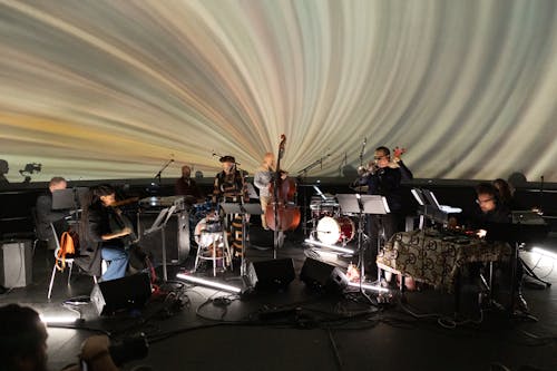 Nine musicians perform on a stage in front of a multicolored video projection.