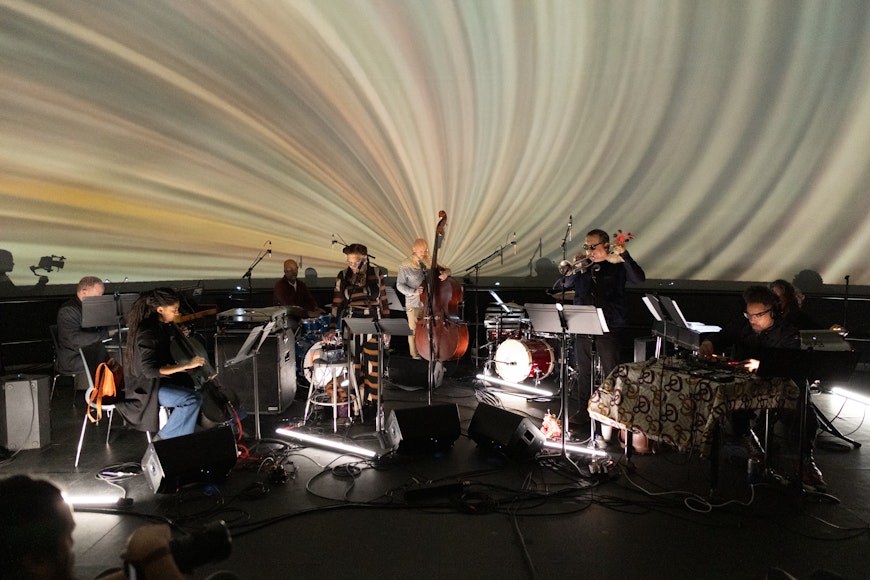 Nine musicians perform on a stage in front of a multicolored video projection.