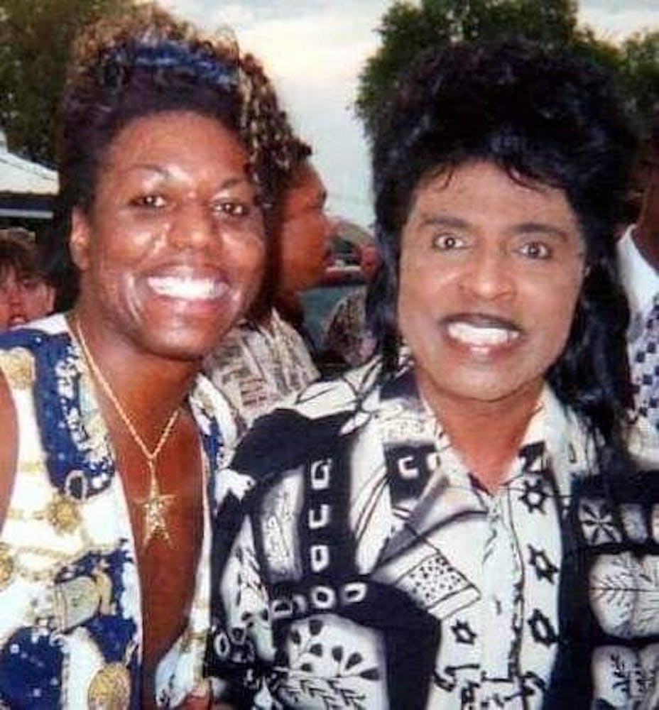 Two men in loud shirts with a pencil mustaches stand next to one another while smiling at the viewer.