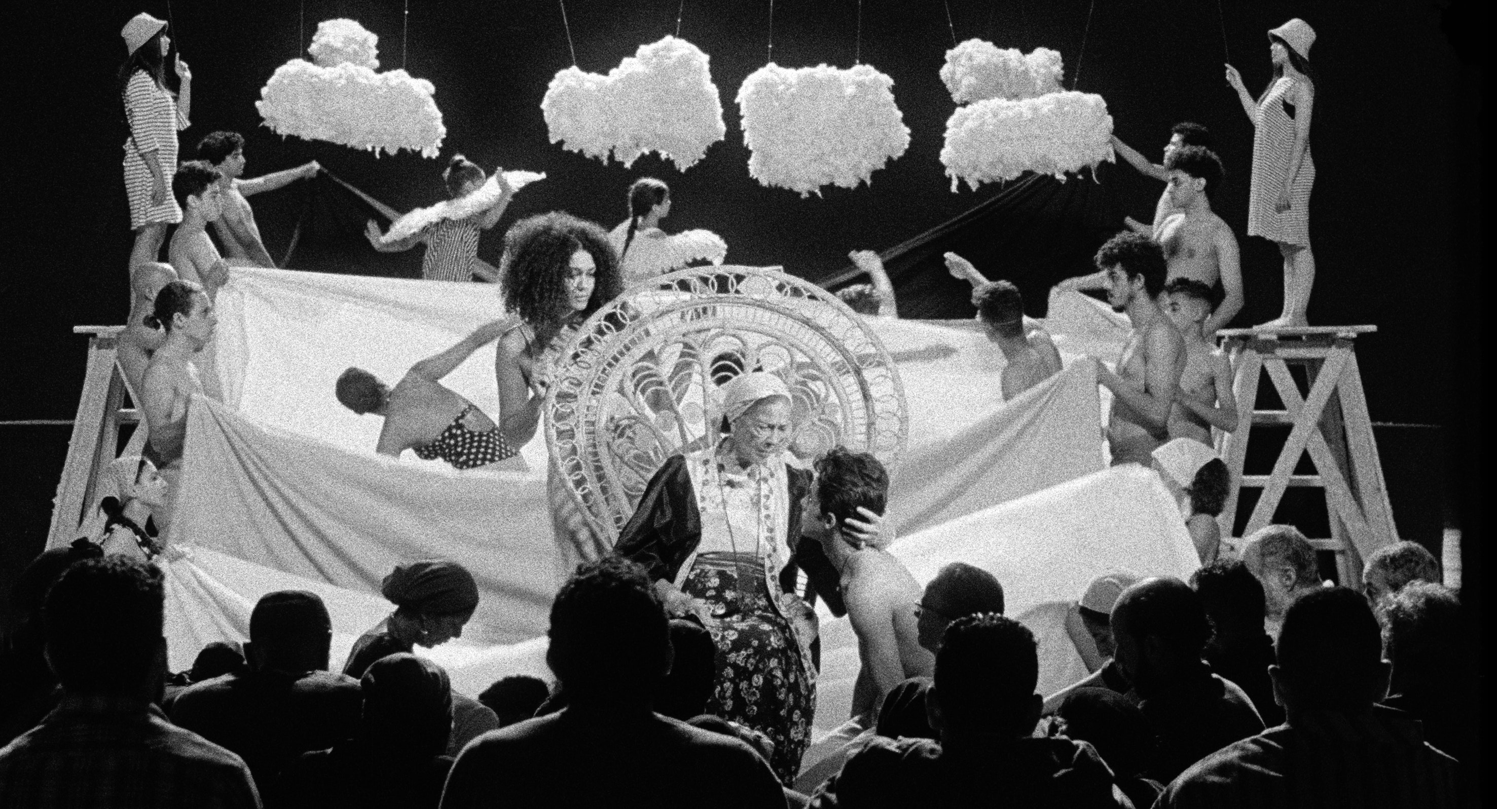 Black and white still of a stage production with an elderly woman at the front center, people holding waves of cloth, and hanging clouds in the back.