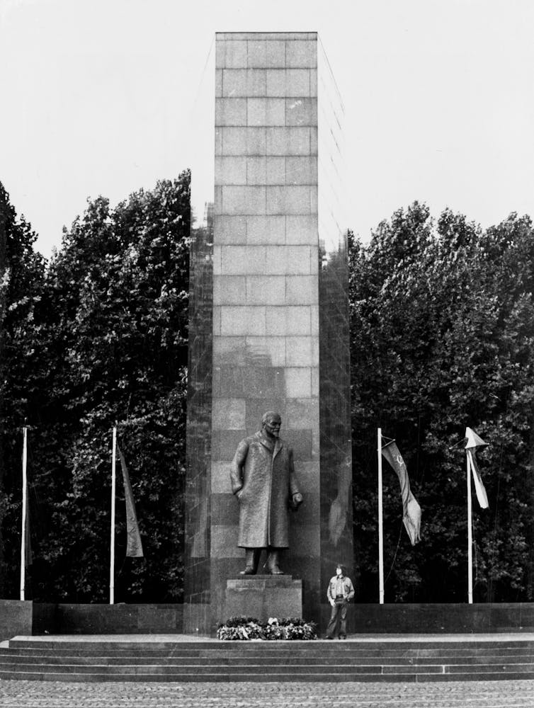 Black and white photo of a man standing next to a large soviet statue of a man outside.
