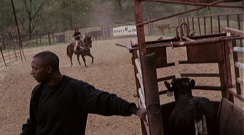 A man in a black sweatshirt leans against a cow pen in a rodeo arena. In the background someone is riding a horse.