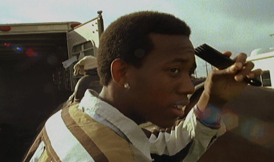 A close up of a young man in a striped shirt holding an afro pick toward his head. There are trailers in the background.
