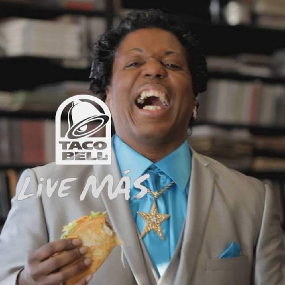 A man in a brightly colored suit with a pencil mustach smiles at the viewer while holding a taco with the Taco Bell logo and tag line 