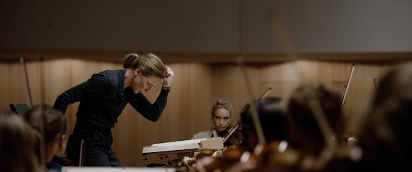 A woman conductor leans forward as she conducts an orchestra.
