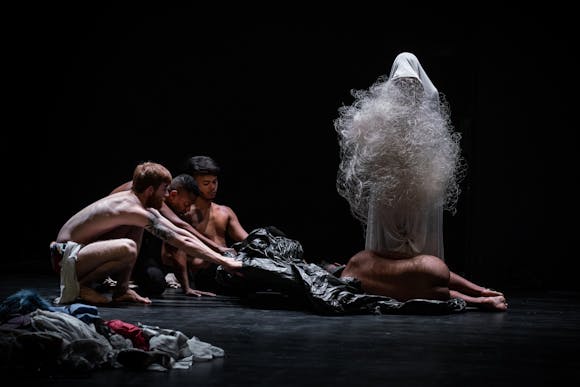 Two adults pull a sleepbag with a nude person on top of it across a stage in front of a figure in while rob and plastic netting.