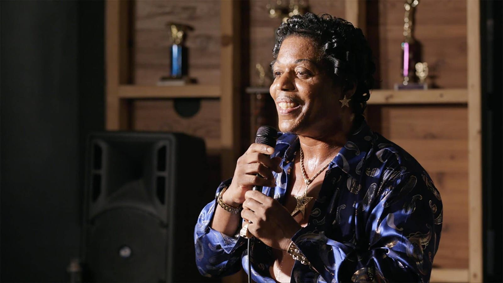 A man in a brightly colored shirt with a pencil mustach does stand up comedy on a stage.