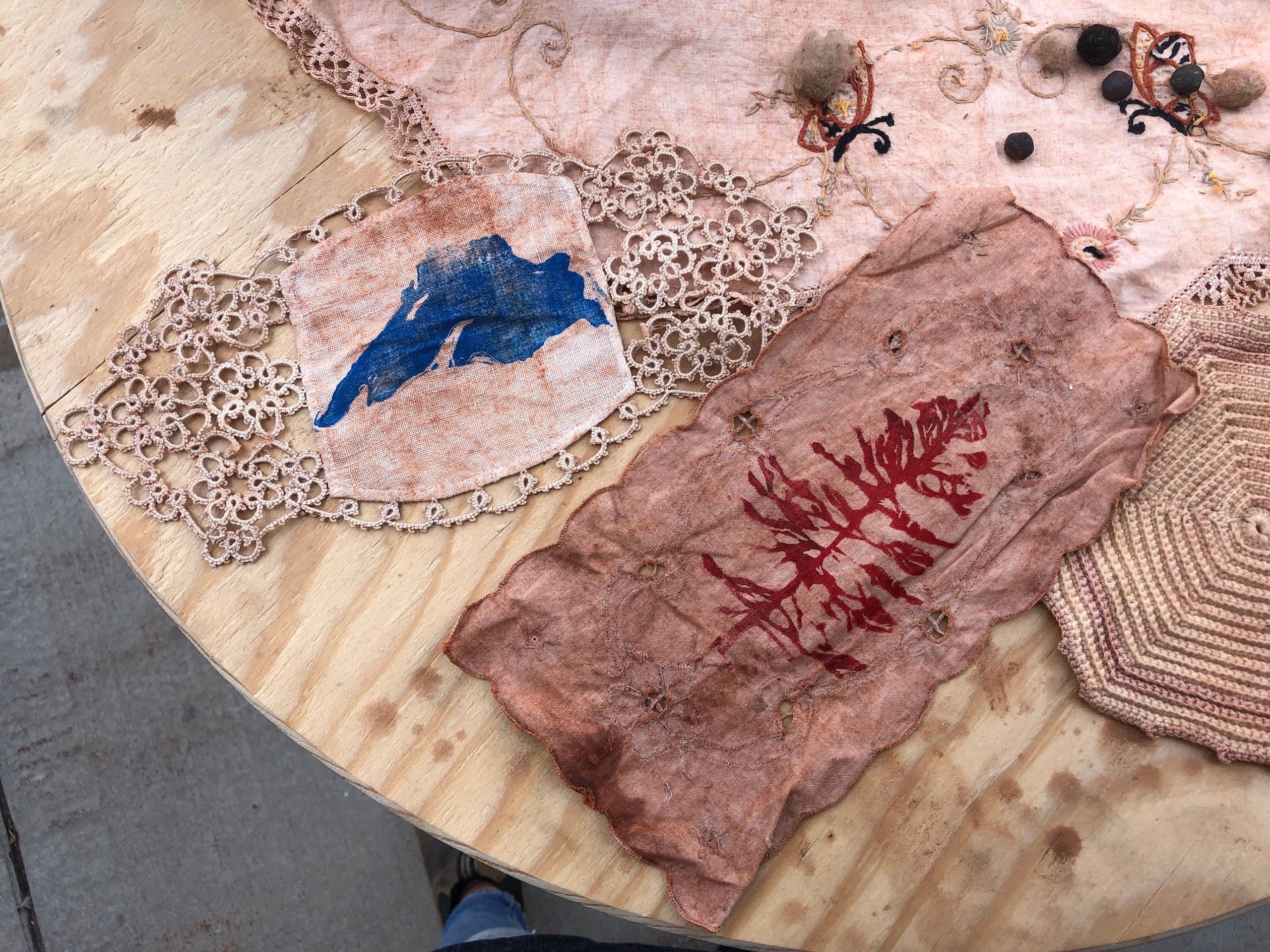Pieces of fabric and embroidery are laid out on a wooden table, stained with red dirt and printed with a red tree and a blue outline of Lake Superior.