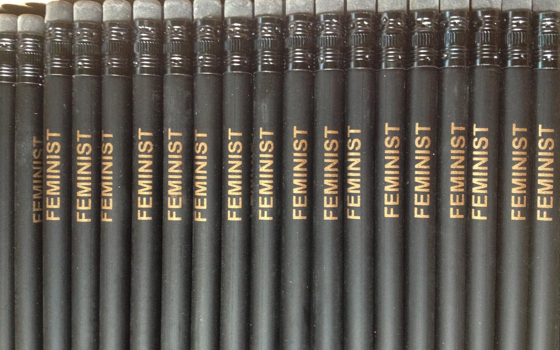 A series of black penicls with the word "feminist" printed on them are lined up on a tabletop.