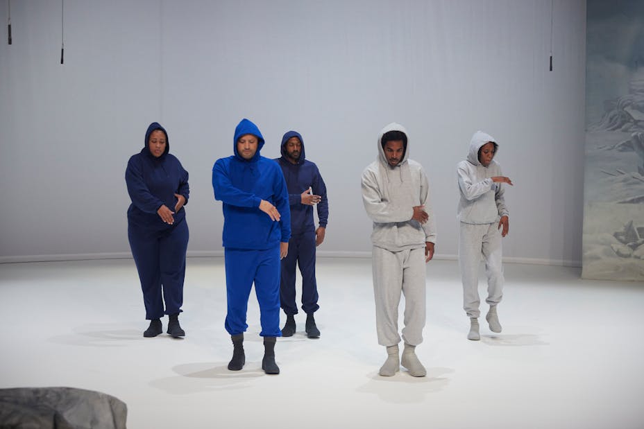 Five dancers wearing hoodies and sweatpants of varying colors move within a white space.