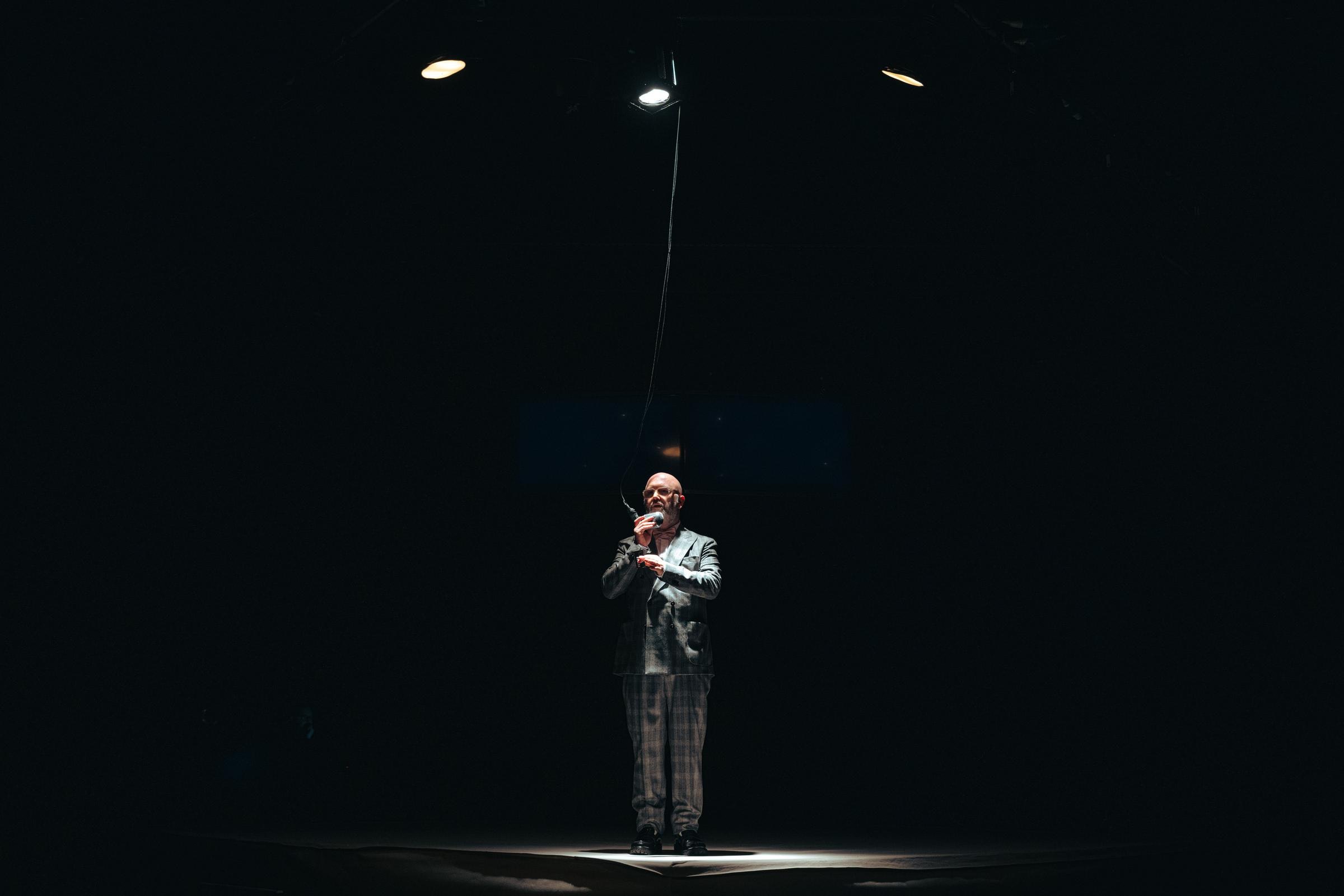 A performer with light skin speaks into a hanging microphone.