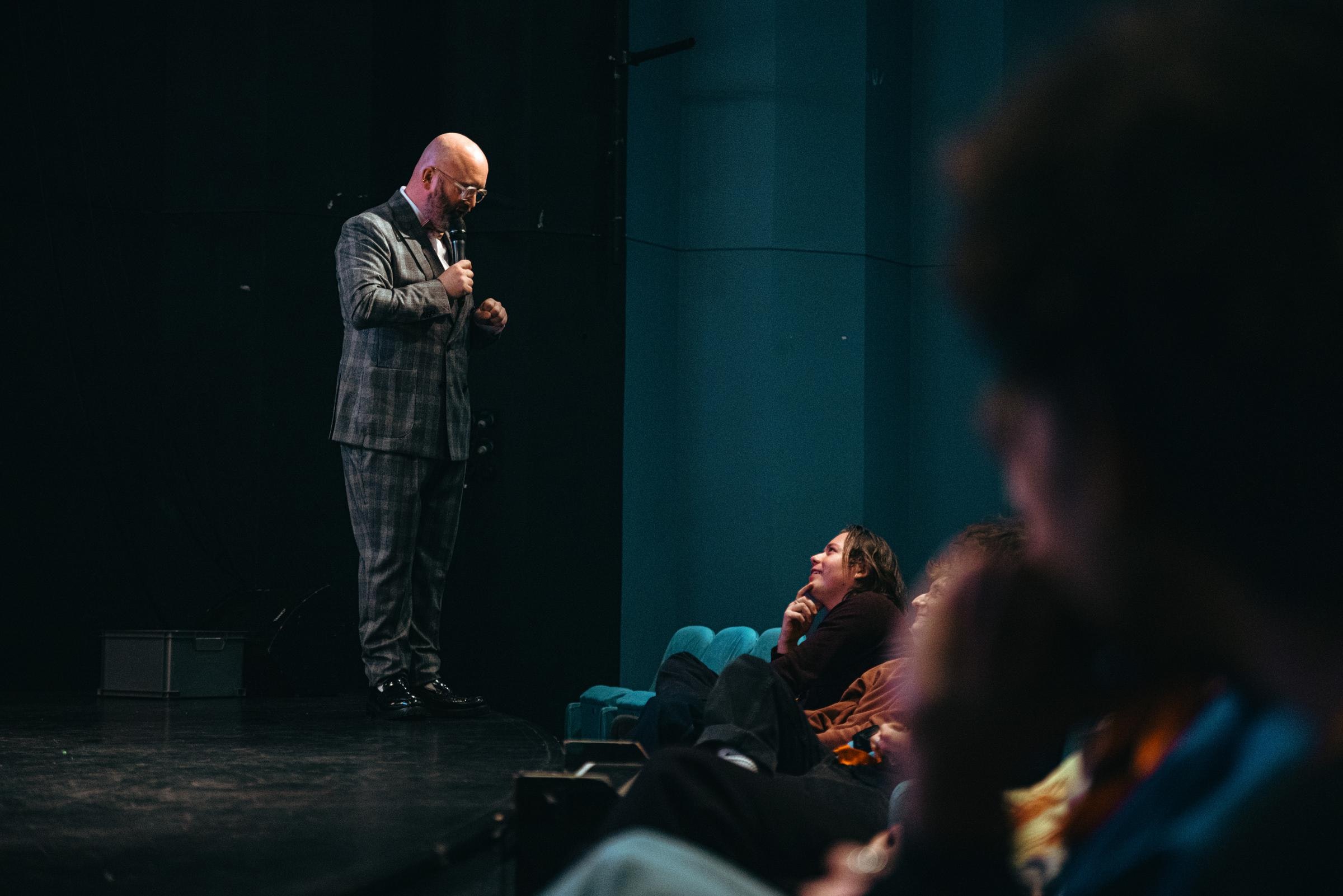 A performer with light skin with a microphone speaks with an audience member at the end of the stage.