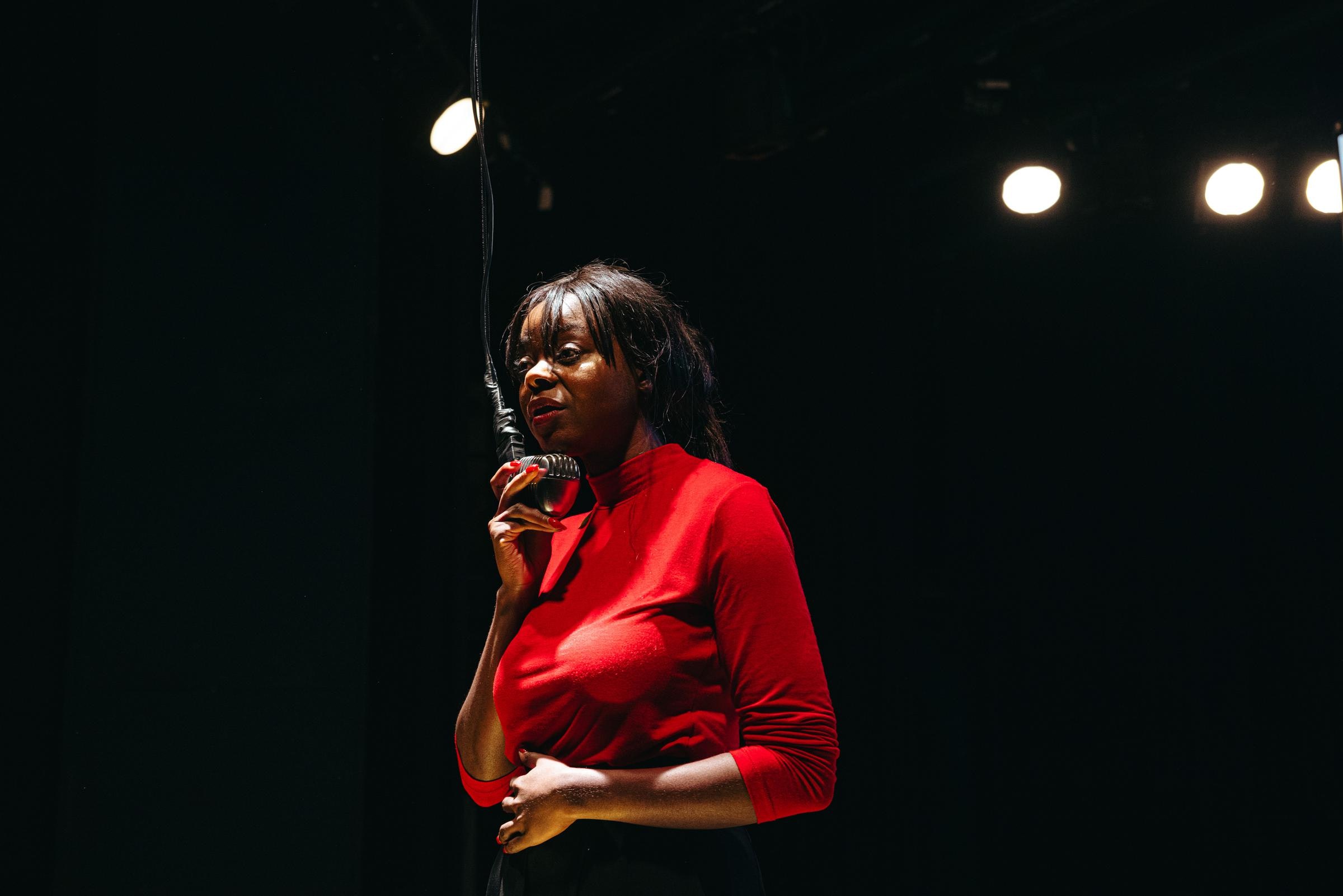 A performer with dark skin speaks into a hanging microphone.