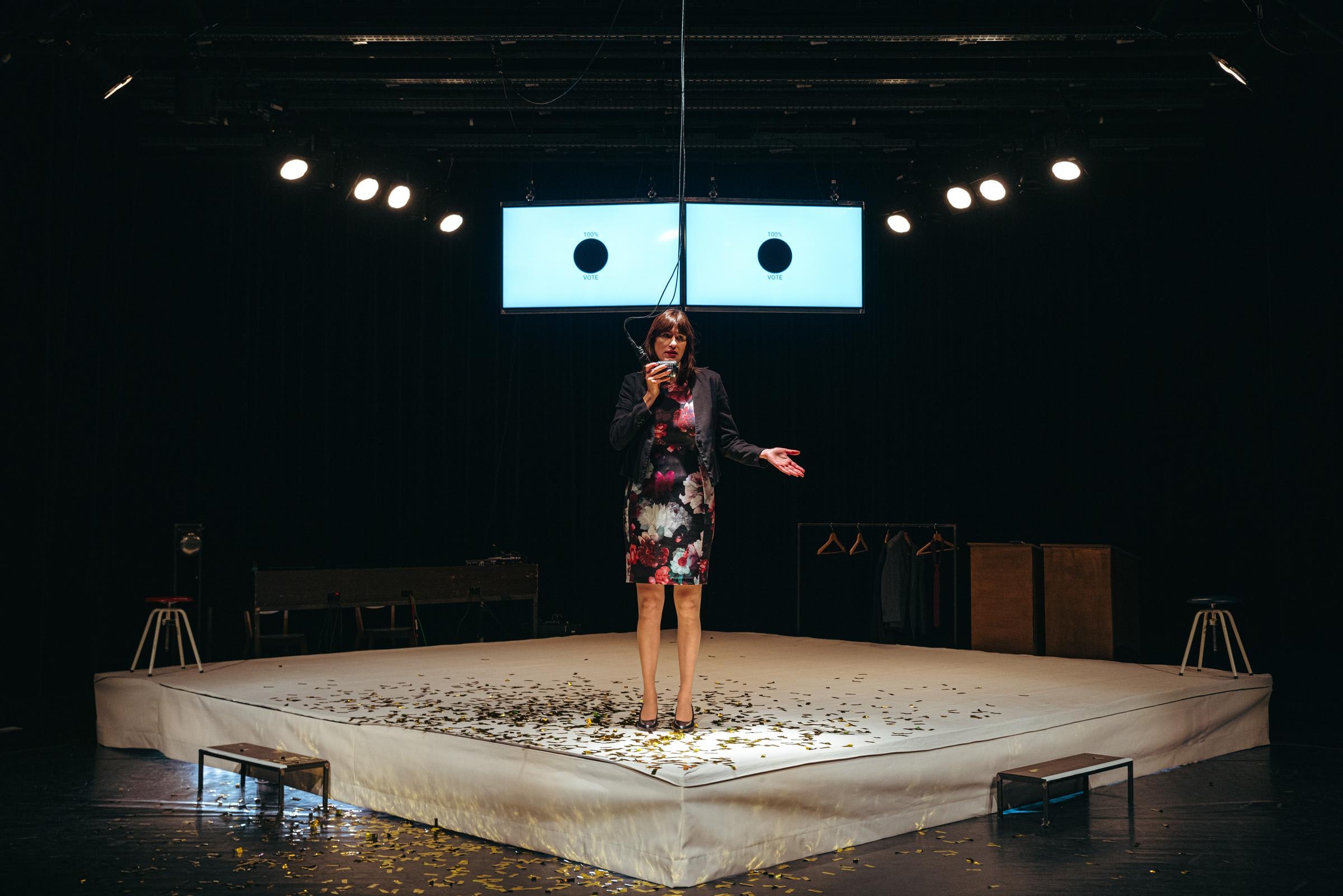 A performer with light skin stands on a raised platform speaking into a hanging microphone.