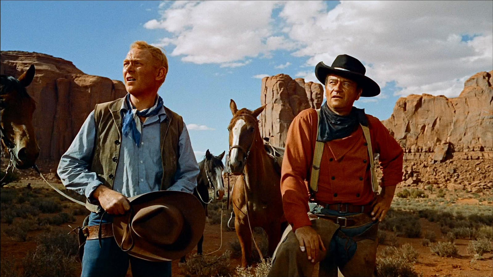 Two men squint looking upward, in a desert landscape with tall red rock formations. They wear western clothing and stand with three horses.