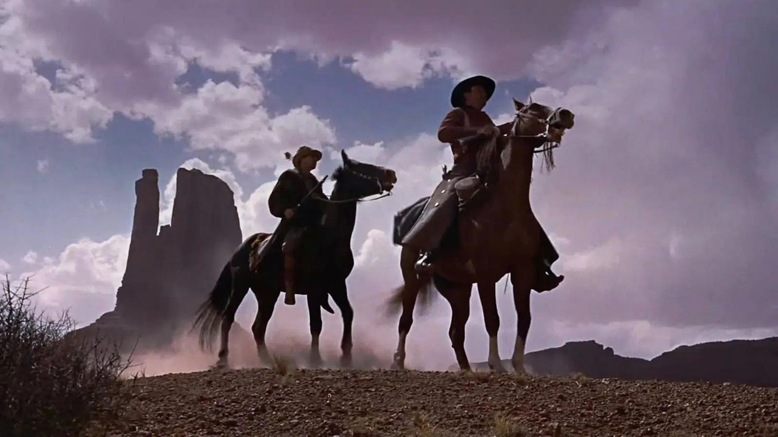 Still image looking up toward two men on horseback in a rocky western landscape, tall red rock formations in the distance against a blue cloudy sky.