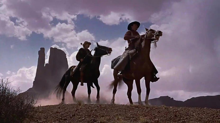 Still image looking up toward two men on horseback in a rocky western landscape, tall red rock formations in the distance against a blue cloudy sky.
