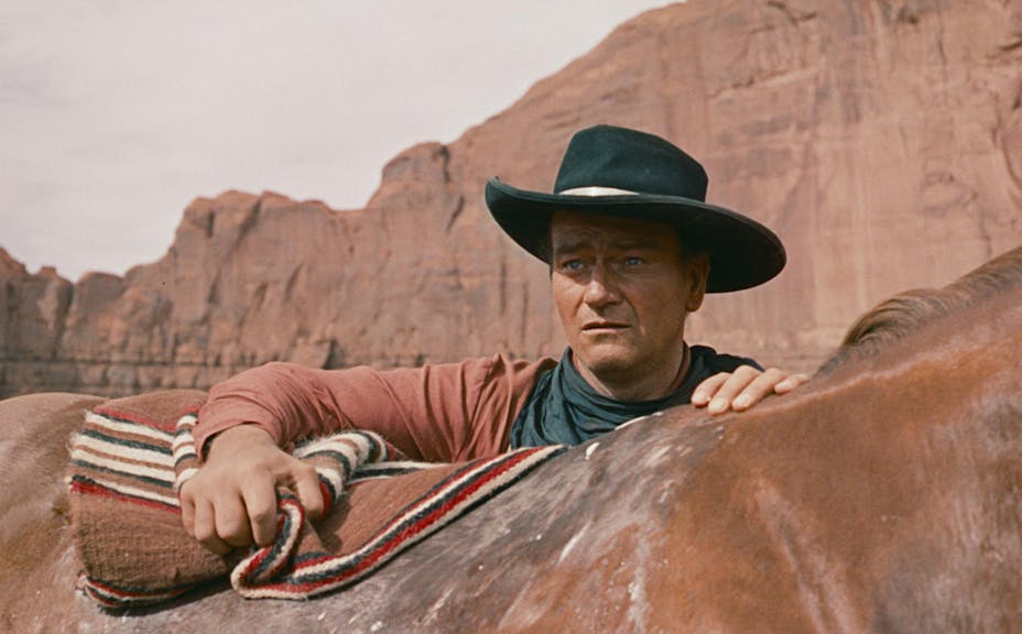 John Wayne in a red rocky western landscape, wears a black hat and puts a blanket on the back of a brown horse while he looks out in the distance.