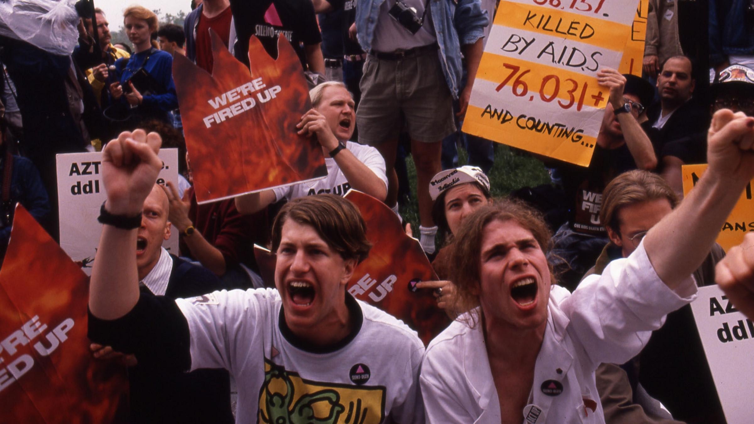 A crowd of protestors raise their fists and shout, holding signs about AIDS medications and death tolls.
