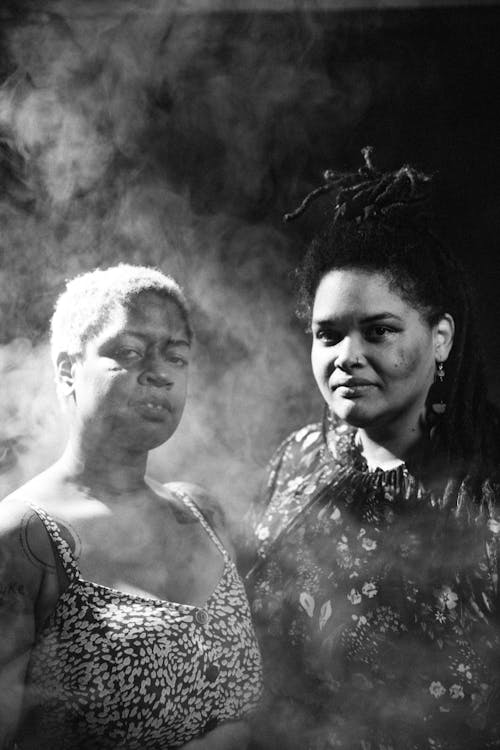 Two artists look into the camera standing in a balck space filled with smoke.