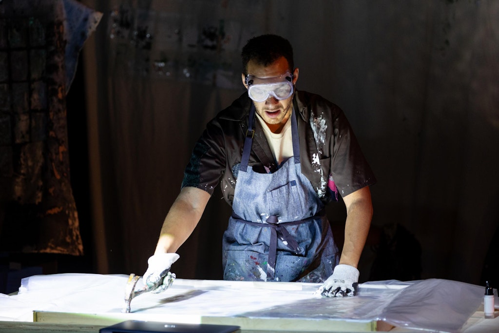 A performer with medium dark skin hammers a canvas while wearing clear goggles.