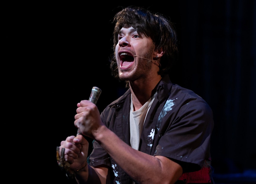 A performer with medium dark skin holds a hammer like a microphone.