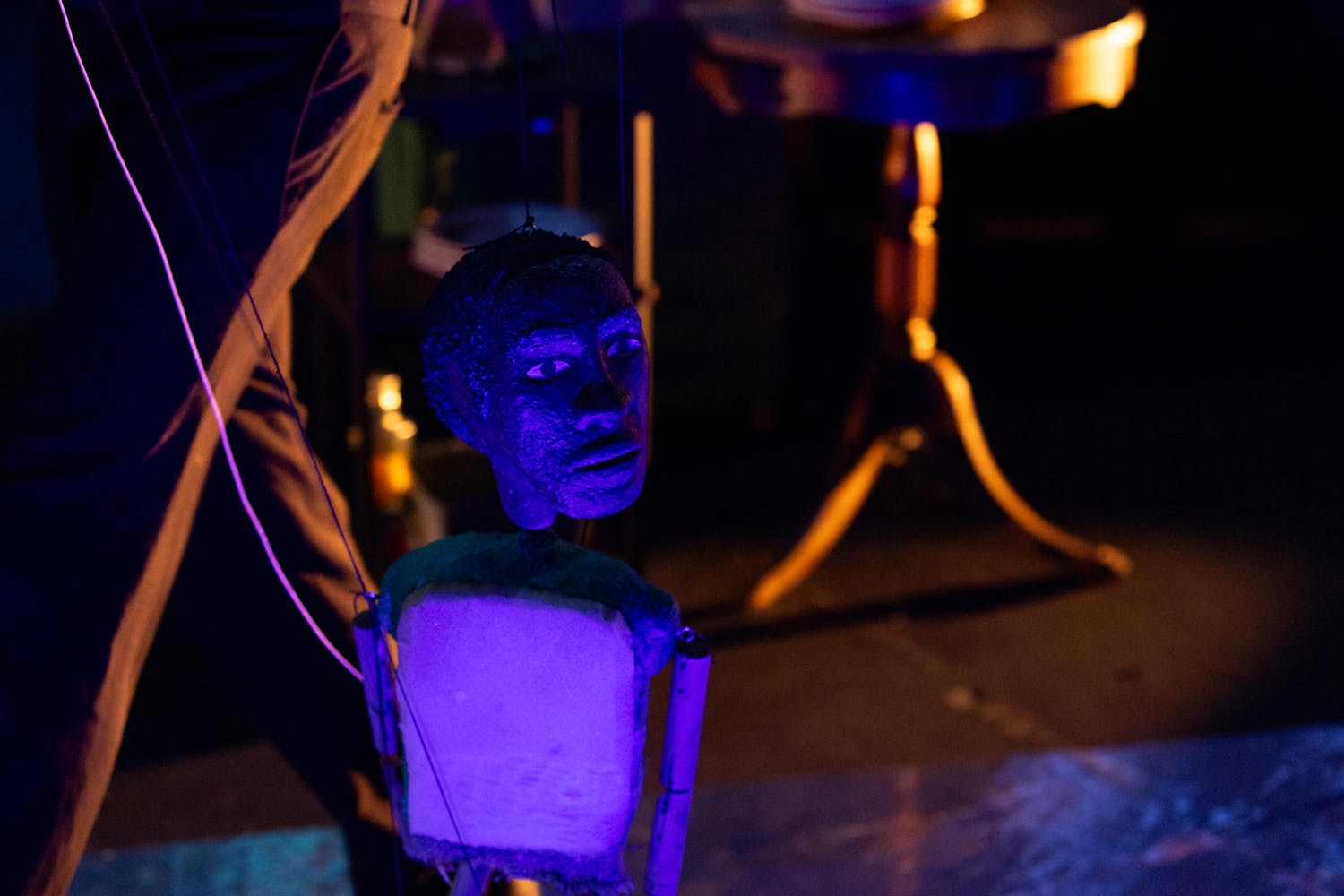 A puppet with dark skin stands in front of a wooden chair and table.