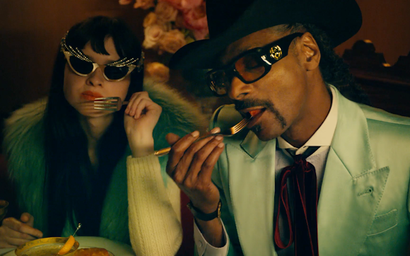 Snoop Dogg wearing sunglasses, a cowboy hat and a mint green suit eats at a table with a woman in sunglasses and a fuzzy coat.