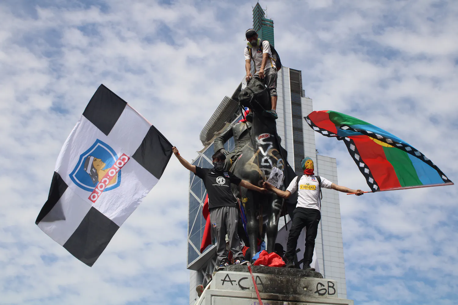 A small group of adults stand atop a outdoor sculpture of a man on a horse while waving flags and protesting.