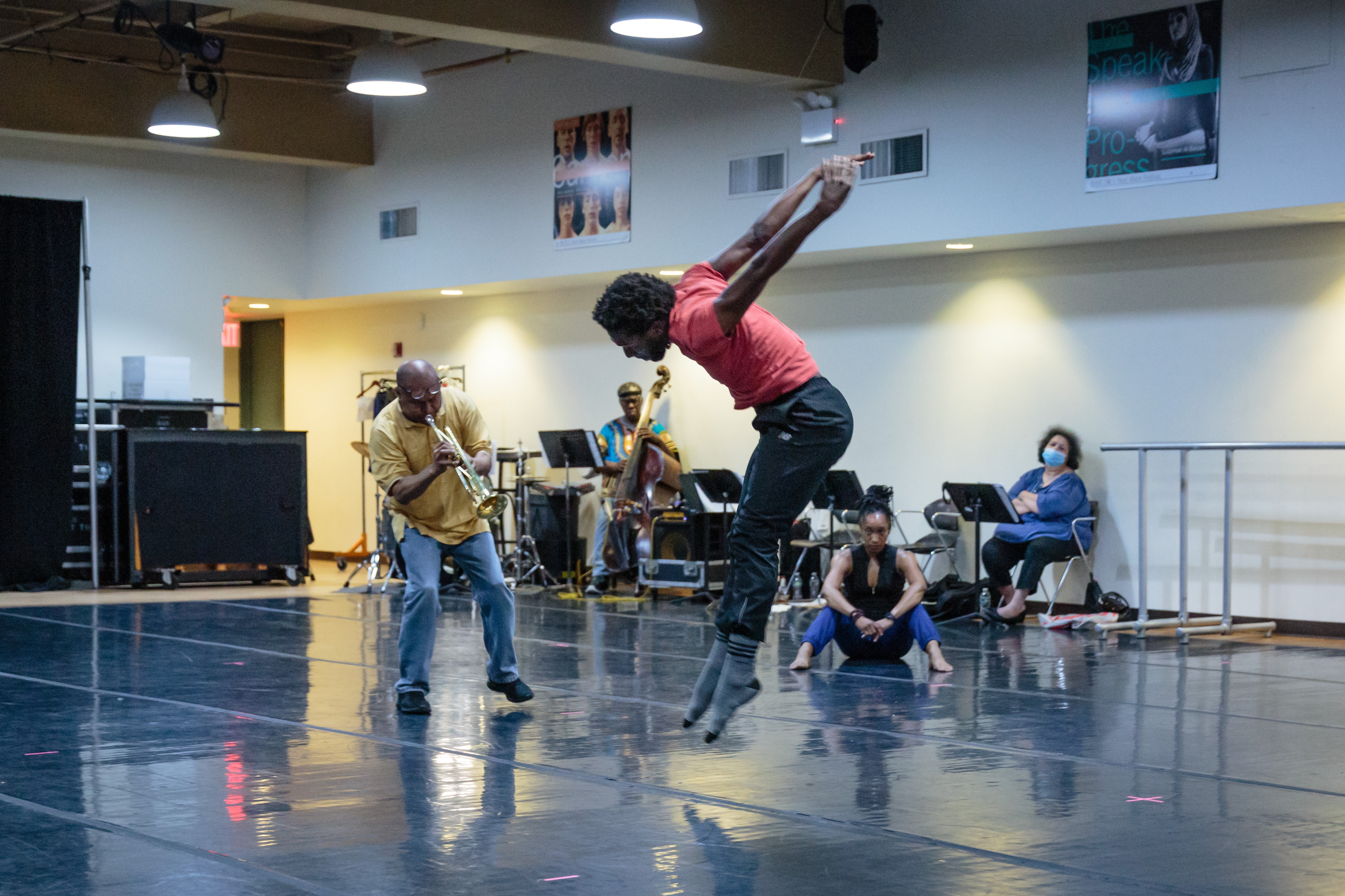A dancer with dark skin jumps up while an adult with dark skin plays the trumpet in a dancer space.