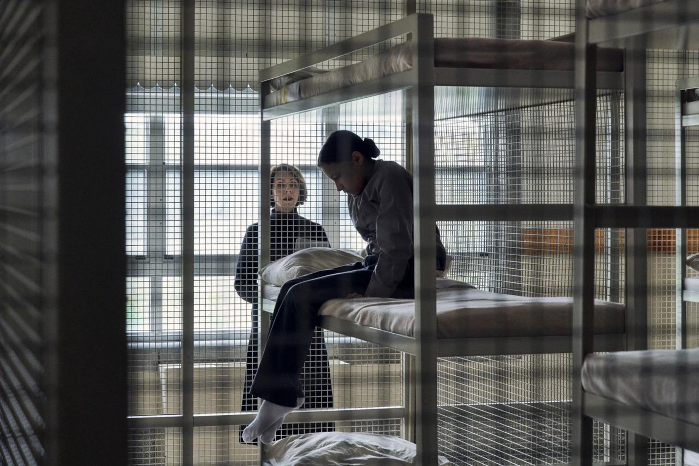 A woman talks to another women who is sitting on a bunkbed in a jail cell.