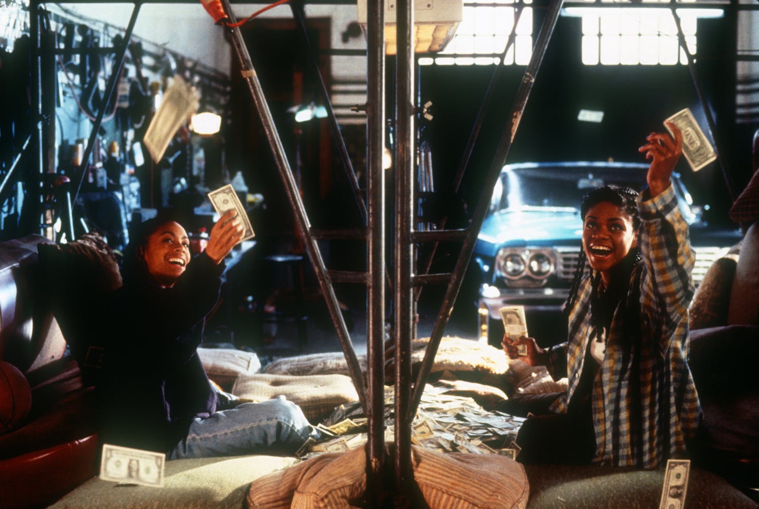 Two women sitting on the floor of a auto mechanic garage laugh and throw money into the air.