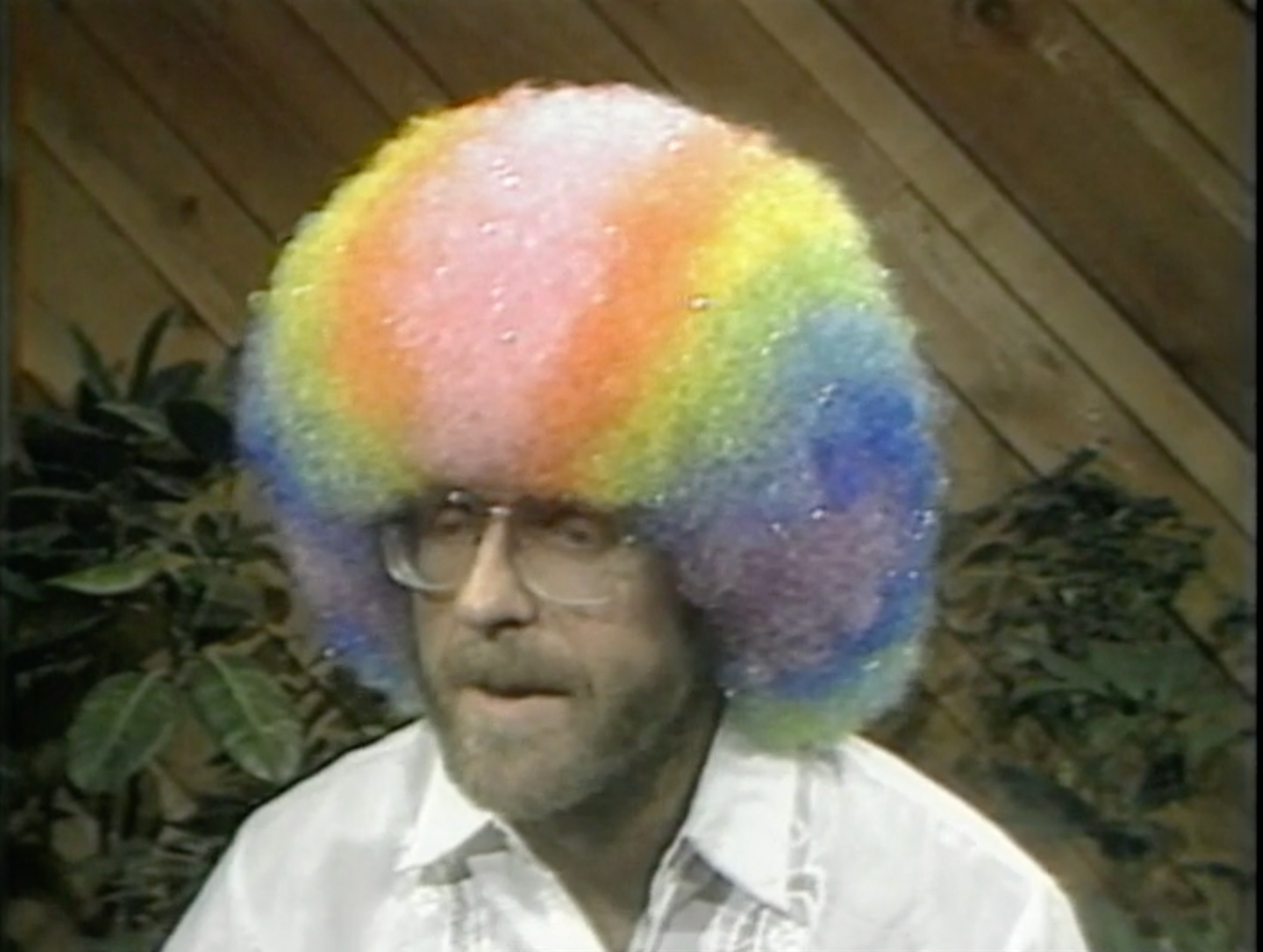 A man with glasses wears a rainbow colored afro wig while sitting in a shair and talking to someone unseen.