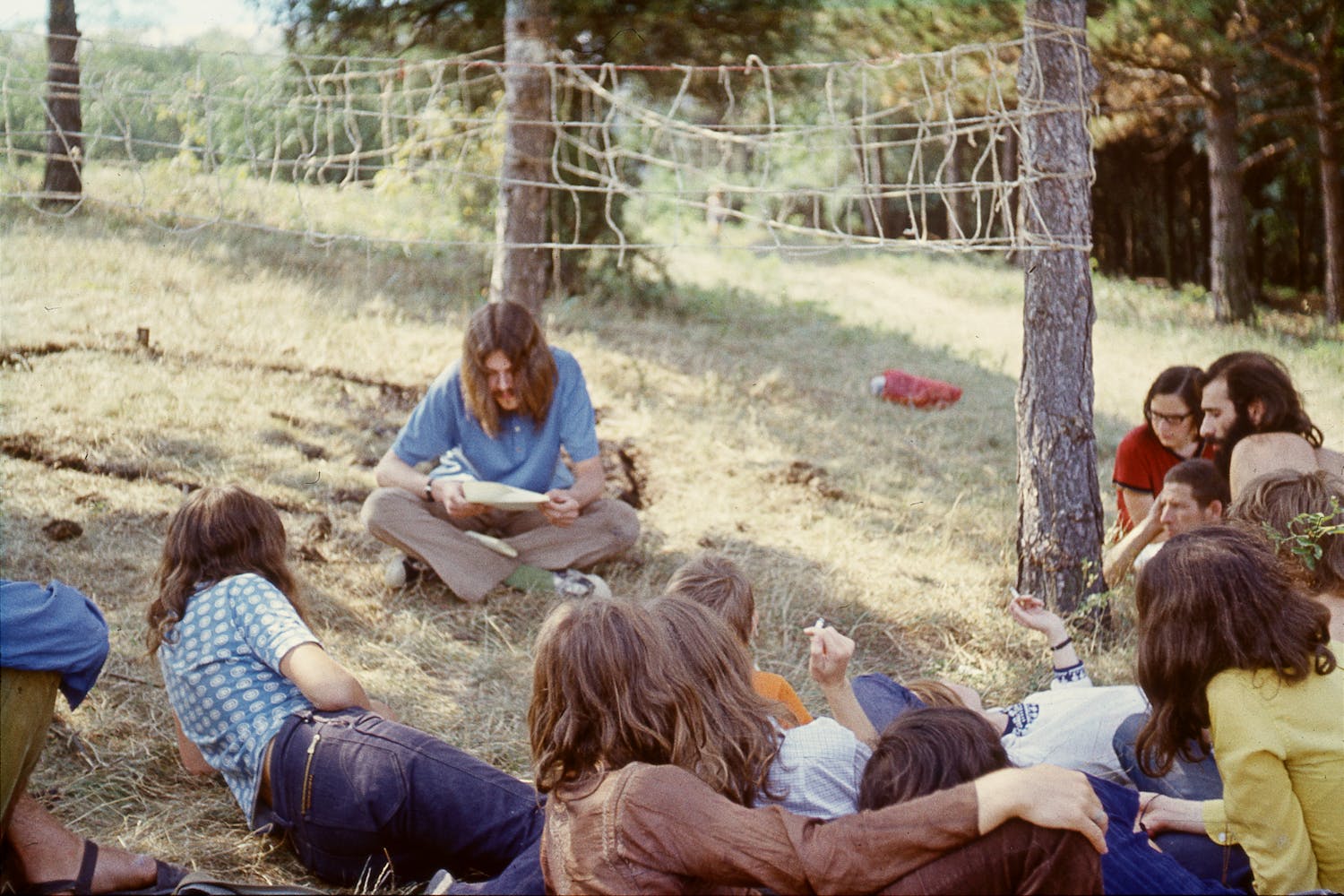 A ground of adults with long hair sit outside on the grass with rope woven between trees behind them.