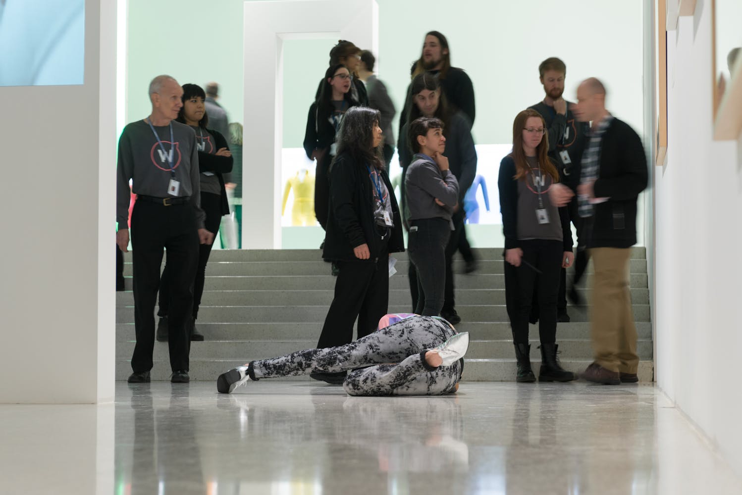 Adults in a white gallery watch a performer on the floor.