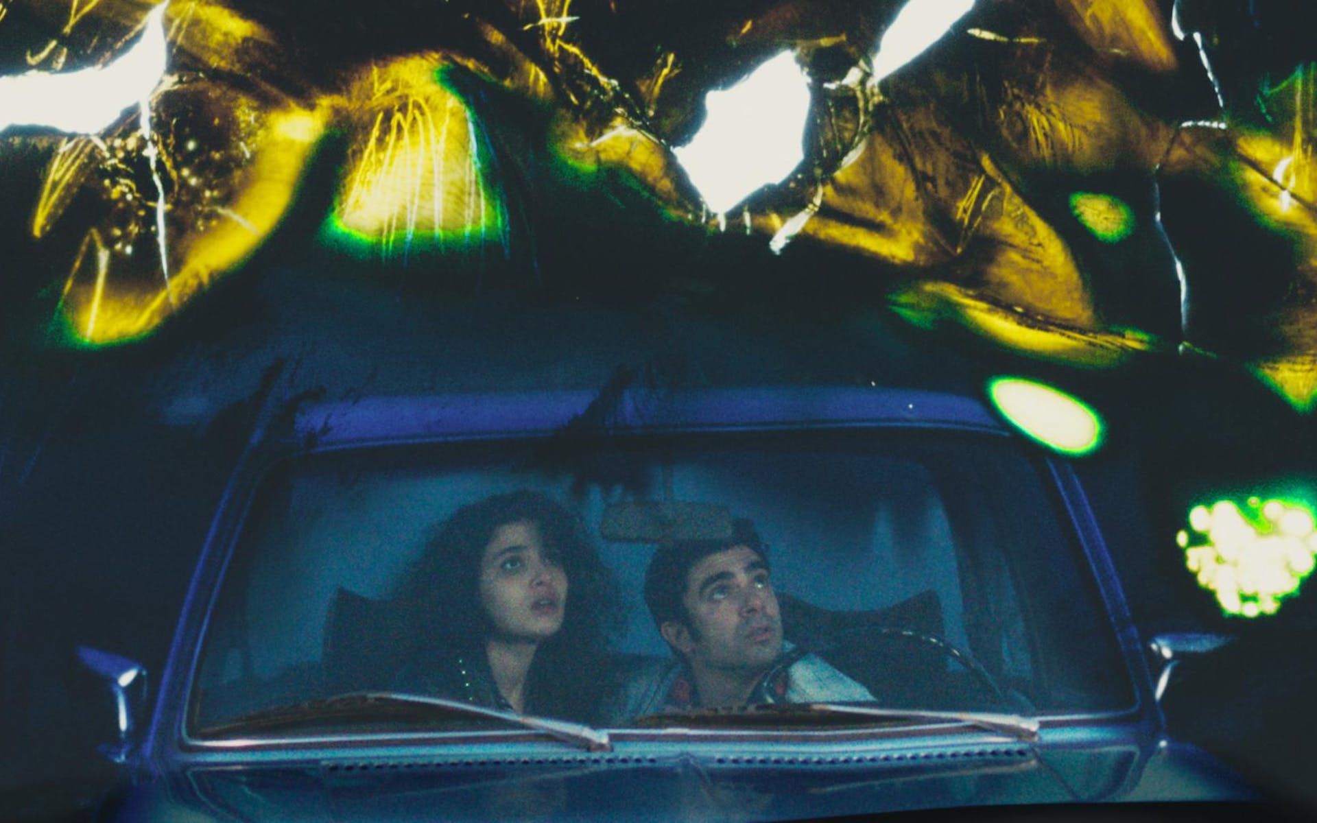 A man and a woman sit a car and look up at strange lights in the sky.