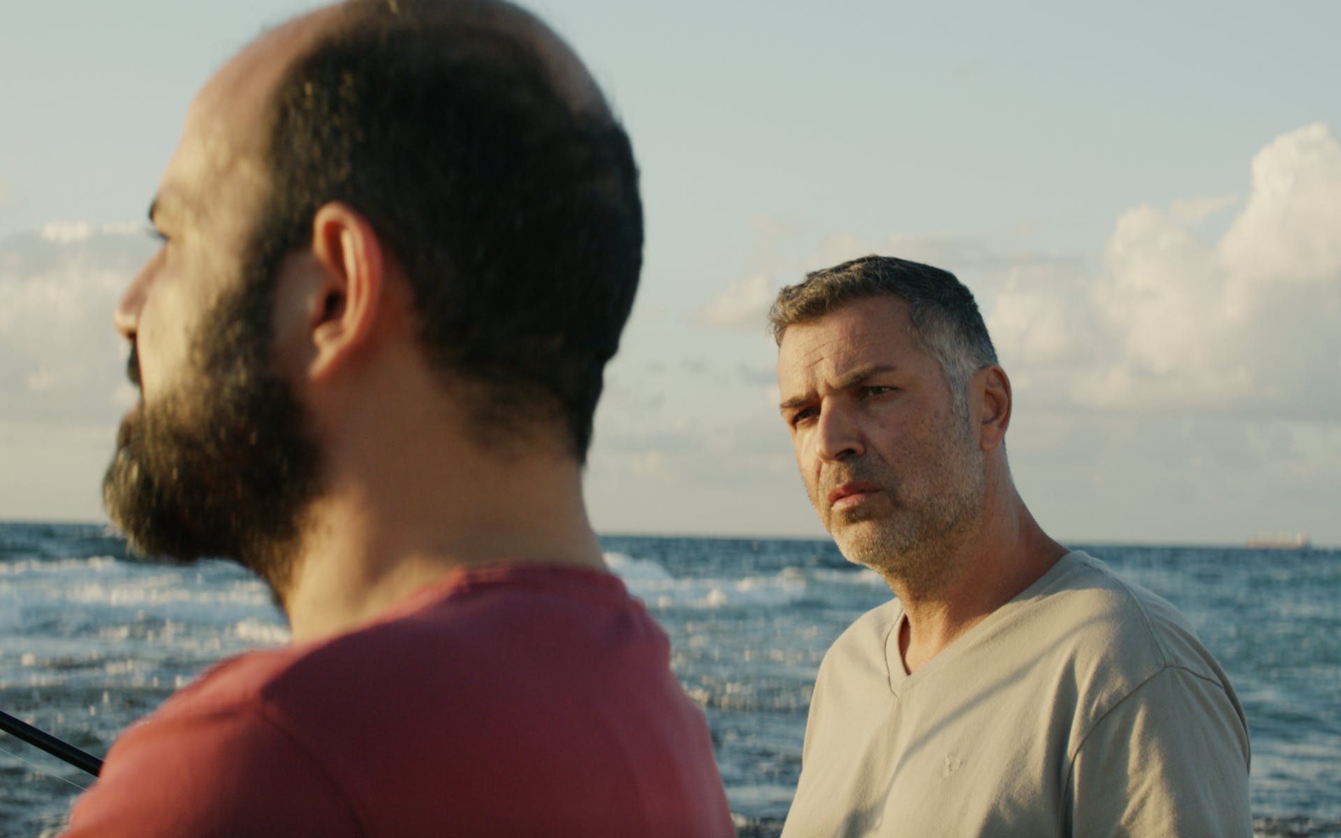 A man in a gray shirt looks at another slightly blurred man in the foreground who looks out at the sea and clouds beyond them.