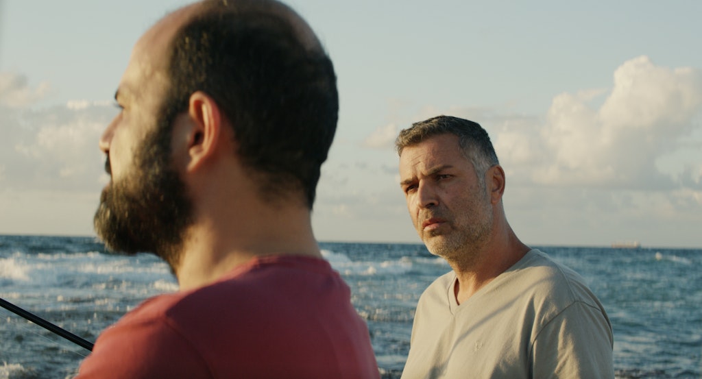 A man in a gray shirt looks at another slightly blurred man in the foreground who looks out at the sea and clouds beyond them.