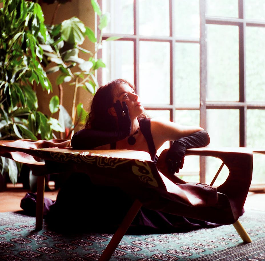 An adult with light skin and long brown hair is sitting in a wooden chair wears long gloves and looks up