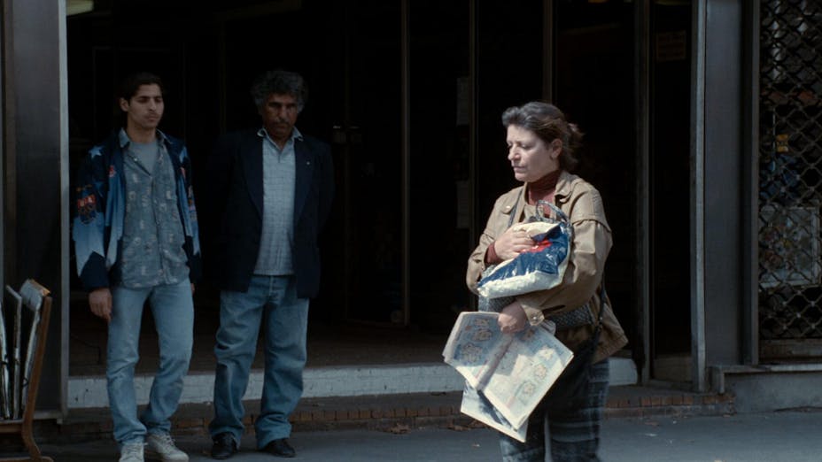 A woman on the street carrying bags and newspapers. Two men are walking in her direction in the background.