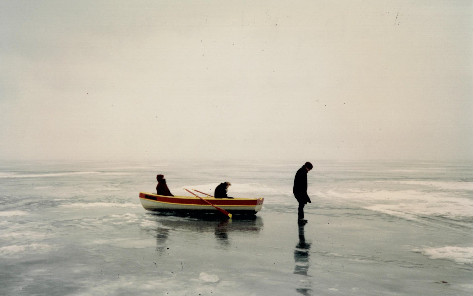 Two figures sit in a rowboat on top of a frozen lake, and a third stands on the ice. Gray sky and ice make up most of the image.
