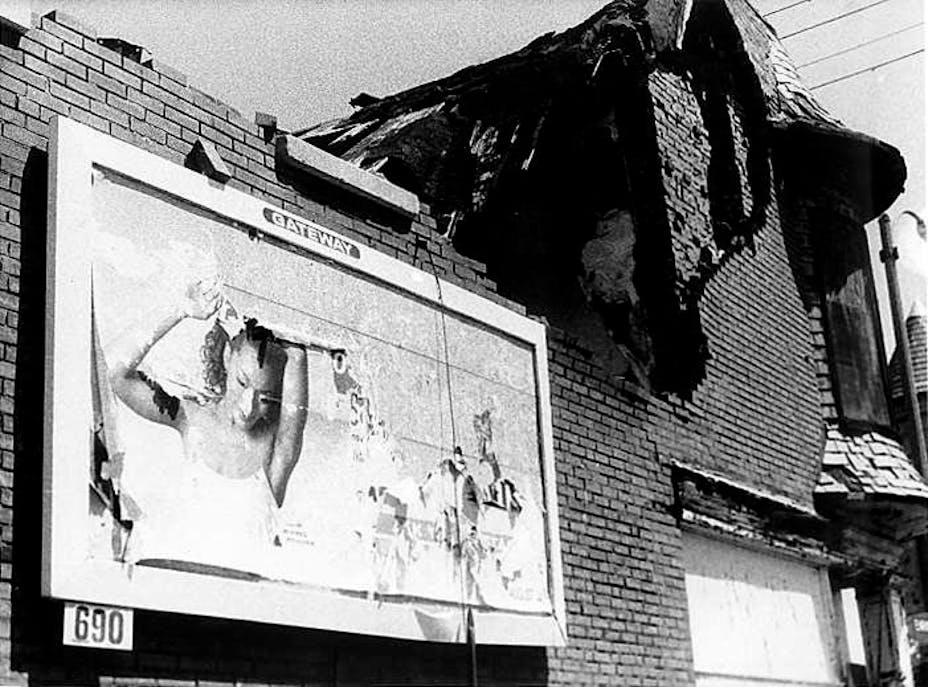 Black and white film still of a partially fallen-down brick building. There is a billboard on it that is peeling off.