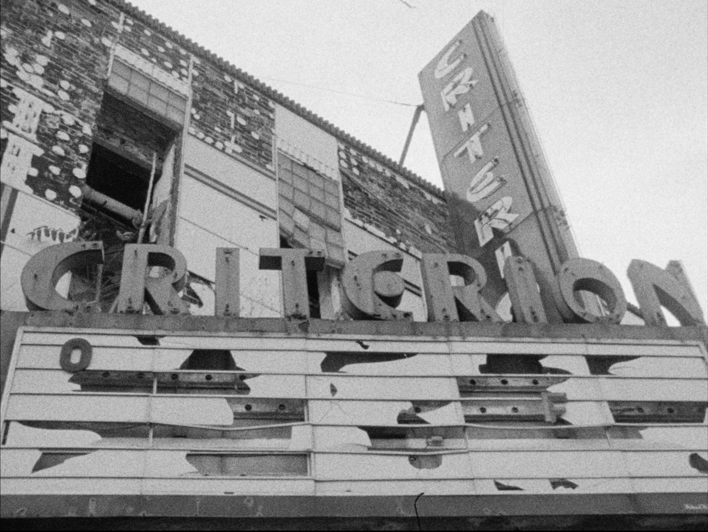Black and white film still of a decaying theater marquee that reads CRITERION. The building facade and windows are deteriorated.
