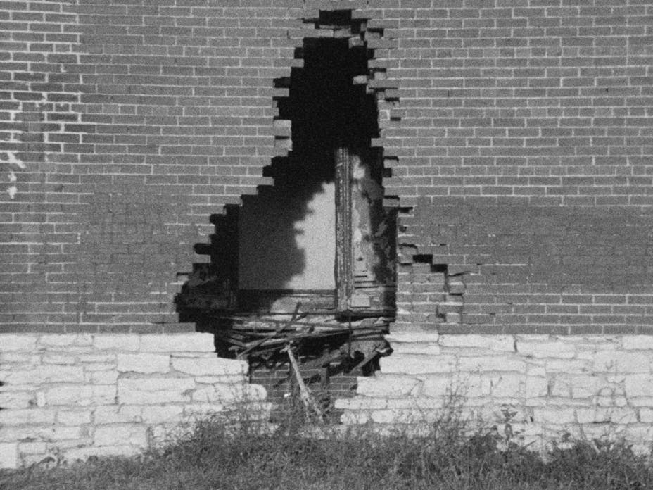 Black and white film still of a brick wall with a large hole broken through it. Through the hole are parts of building materials.