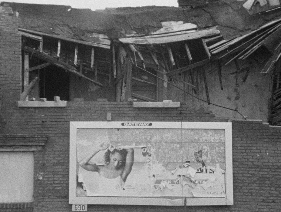 Black and white film still of a partially fallen-down brick building. There is a billboard on it that is peeling off.