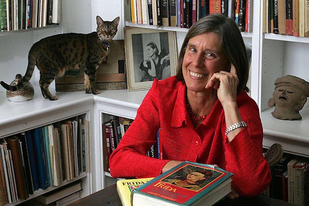 An adult with light skin, wearing a red shirt and silver watch, has medium legth brown hair and smiles at the viewer with a hand to their face. A cat stands on a bookshelf behind.